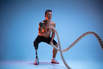 Image showing Athlete with disabilities or amputee isolated on blue studio background. Professional male sportsman with leg prosthesis training with weights in neon