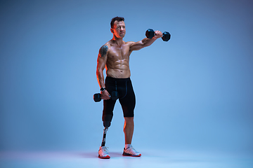 Image showing Athlete with disabilities or amputee isolated on blue studio background. Professional male sportsman with leg prosthesis training with weights in neon