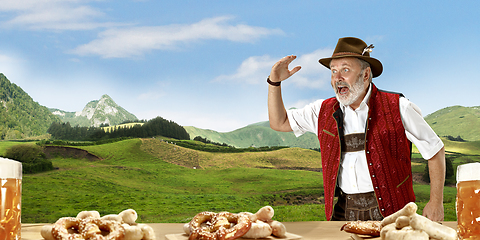 Image showing The senior happy smiling man with beer dressed in traditional Austrian or Bavarian costume holding mug of beer, mountains on background, flyer