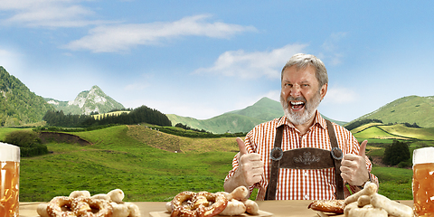 Image showing The senior happy smiling man with beer dressed in traditional Austrian or Bavarian costume holding mug of beer, mountains on background, flyer