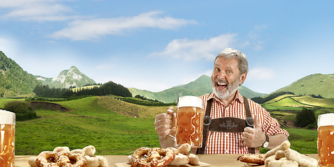 Image showing The senior happy smiling man with beer dressed in traditional Austrian or Bavarian costume holding mug of beer, mountains on background, flyer