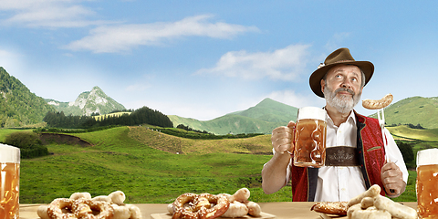 Image showing The senior happy smiling man with beer dressed in traditional Austrian or Bavarian costume holding mug of beer, mountains on background, flyer