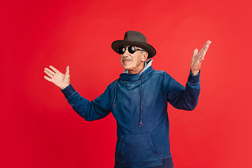 Image showing Senior man in stylish eyewear and hat isolated on red background. Tech and joyful elderly lifestyle concept