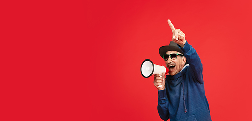 Image showing Senior man in stylish eyewear and hat isolated on red background. Tech and joyful elderly lifestyle concept