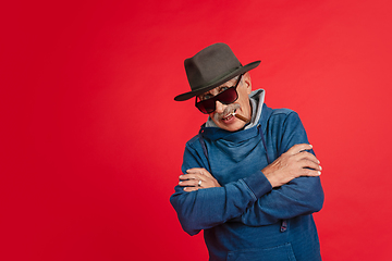 Image showing Senior man in stylish eyewear and hat isolated on red background. Tech and joyful elderly lifestyle concept