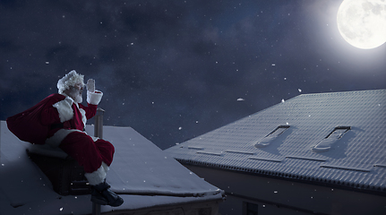 Image showing Emotional Santa Claus congratulating with New Year and Christmas, sitting on roof of the house in midnight with full moon