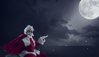 Image showing Emotional Santa Claus congratulating with New Year and Christmas, sitting on roof of the house in midnight with full moon