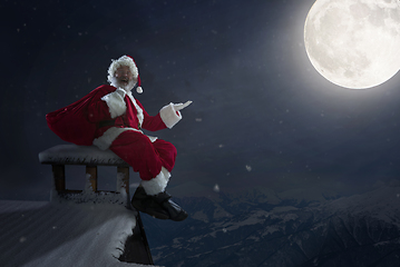 Image showing Emotional Santa Claus congratulating with New Year and Christmas, sitting on roof of the house in midnight with full moon