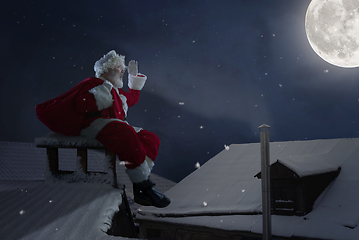 Image showing Emotional Santa Claus congratulating with New Year and Christmas, sitting on roof of the house in midnight with full moon