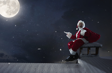 Image showing Emotional Santa Claus congratulating with New Year and Christmas, sitting on roof of the house in midnight with full moon