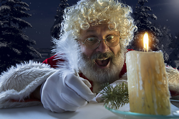 Image showing Emotional Santa Claus congratulating with New Year and Christmas, writing a letter, wish list in midnight with candle