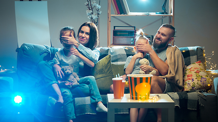 Image showing Happy family watching projector, TV, movies with popcorn in the evening at home. Mother, father and kids spending time together.