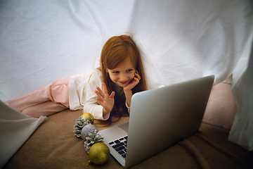 Image showing Happy caucasian little girl during video call or messaging with Santa using laptop and home devices, looks delighted and happy