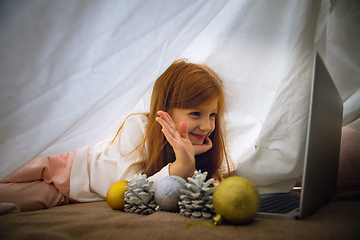 Image showing Happy caucasian little girl during video call or messaging with Santa using laptop and home devices, looks delighted and happy