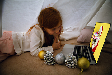 Image showing Happy caucasian little girl during video call or messaging with Santa using laptop and home devices, looks delighted and happy
