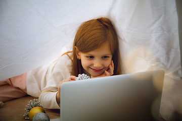 Image showing Happy caucasian little girl during video call or messaging with Santa using laptop and home devices, looks delighted and happy