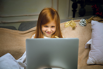 Image showing Happy caucasian little girl during video call or messaging with Santa using laptop and home devices, looks delighted and happy