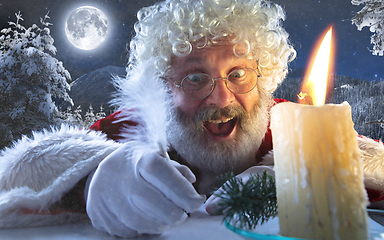 Image showing Close-up portrait of emotional Santa Claus writing a letter,wish list with New Year and Christmas in midnight with candle