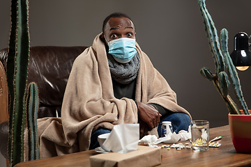 Image showing Young man wrapped in a plaid looks sick, ill, sneezing and coughing sitting at home indoors in face mask.