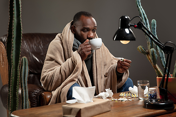 Image showing Young man wrapped in a plaid looks sick, ill, sneezing and coughing sitting at home indoors.