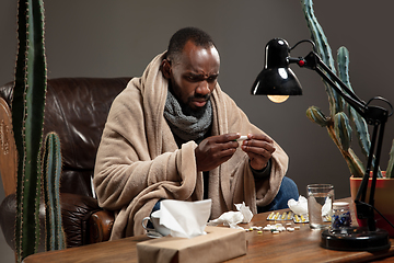 Image showing Young African-american man wrapped in a plaid looks sick, ill, sitting at home indoors. Healthcare concept