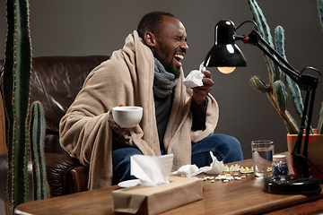 Image showing Young man wrapped in a plaid looks sick, ill, sneezing and coughing sitting at home indoors.