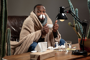 Image showing Young African-american man wrapped in a plaid looks sick, ill sitting at home indoors.