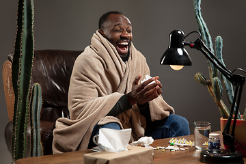 Image showing Young man wrapped in a plaid looks sick, ill, sneezing and coughing sitting at home indoors.