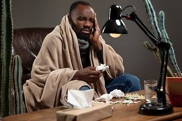 Image showing Young African-american man wrapped in a plaid looks sick, ill, has terrible headache sitting at home indoors.