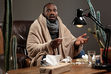 Image showing Young African-american man wrapped in a plaid looks sick, ill, sitting at home indoors. Healthcare concept