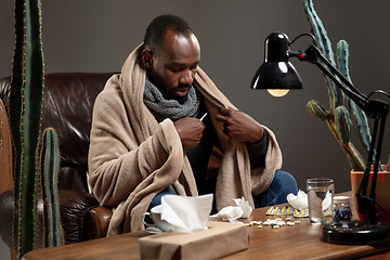 Image showing Young African-american man wrapped in a plaid looks sick, ill, sitting at home indoors. Healthcare concept