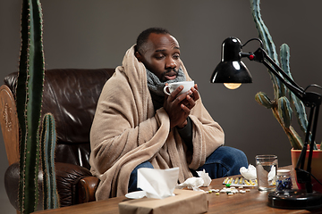 Image showing Young man wrapped in a plaid looks sick, ill, sneezing and coughing sitting at home indoors.