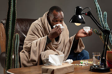 Image showing Young man wrapped in a plaid looks sick, ill, sneezing and coughing sitting at home indoors.