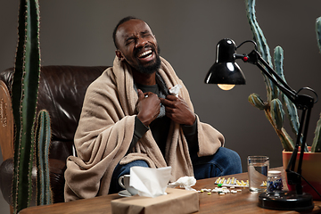 Image showing Young African-american man wrapped in a plaid looks sick, ill, sitting at home indoors. Healthcare concept