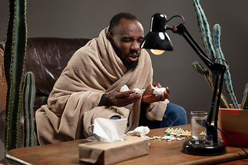 Image showing Young man wrapped in a plaid looks sick, ill, sneezing and coughing sitting at home indoors.