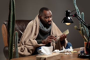 Image showing Young man wrapped in a plaid looks sick, ill, sneezing and coughing sitting at home indoors.