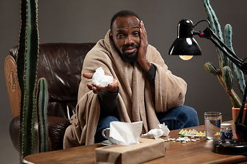 Image showing Young African-american man wrapped in a plaid looks sick, ill, has terrible headache sitting at home indoors.