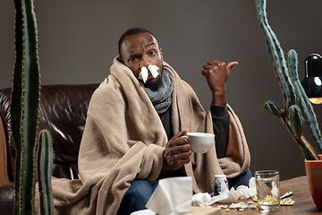 Image showing Young African-american man wrapped in a plaid looks sick, ill sitting at home indoors.