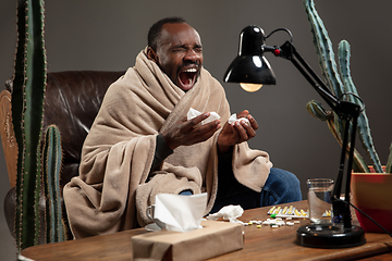 Image showing Young man wrapped in a plaid looks sick, ill, sneezing and coughing sitting at home indoors.