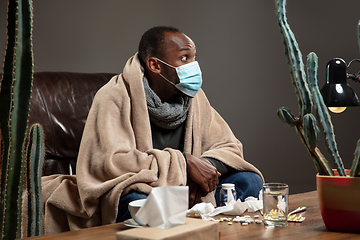 Image showing Young man wrapped in a plaid looks sick, ill, sneezing and coughing sitting at home indoors in face mask.