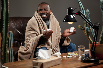 Image showing Young man wrapped in a plaid looks sick, ill, sneezing and coughing sitting at home indoors.