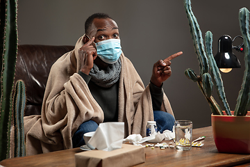 Image showing Young man wrapped in a plaid looks sick, ill, sneezing and coughing sitting at home indoors in face mask.