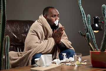 Image showing Young African-american man wrapped in a plaid looks sick, ill sitting at home indoors.