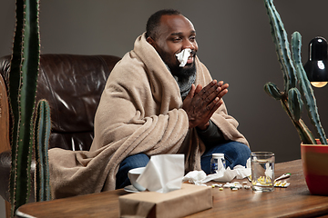 Image showing Young African-american man wrapped in a plaid looks sick, ill sitting at home indoors.