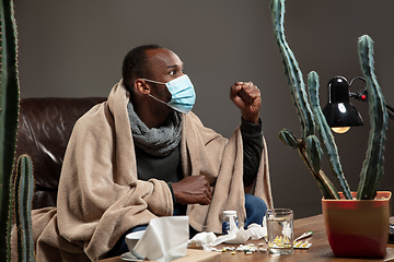 Image showing Young man wrapped in a plaid looks sick, ill, sneezing and coughing sitting at home indoors in face mask.