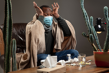 Image showing Young man wrapped in a plaid looks sick, ill, sneezing and coughing sitting at home indoors in face mask.