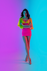 Image showing Beautiful seductive girl in fashionable crimson and yellow outfit on bright gradient pink-blue background in neon light