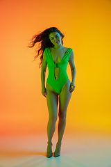Image showing Beautiful seductive girl in fashionable green swimsuit on bright gradient yellow-orange background in neon light