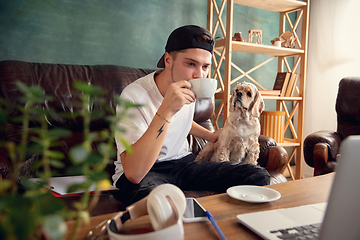 Image showing Young handsome man sitting on sofa at home with his cute dog. Cozy office workplace, remote work, online, e-learning concept.