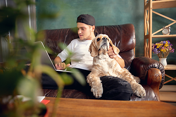 Image showing Young handsome man sitting and working at home with his cute dog. Cozy office workplace, remote work, online learning concept.